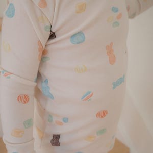 Pode incluir: Um conjunto de pijama rosa claro com gola e punhos brancos. O pijama apresenta um padr&atilde;o de ilustra&ccedil;&otilde;es de doces coloridos, incluindo feij&otilde;es de geleia, ursinhos de goma e doces embalados. A parte superior de manga comprida e as cal&ccedil;as s&atilde;o feitas de um material macio.