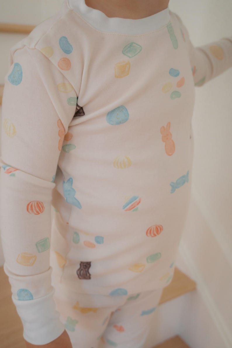 Pode incluir: Um conjunto de pijama rosa claro com gola e punhos brancos. O pijama apresenta um padr&atilde;o de ilustra&ccedil;&otilde;es de doces coloridos, incluindo feij&otilde;es de geleia, ursinhos de goma e doces embalados. A parte superior de manga comprida e as cal&ccedil;as s&atilde;o feitas de um material macio.