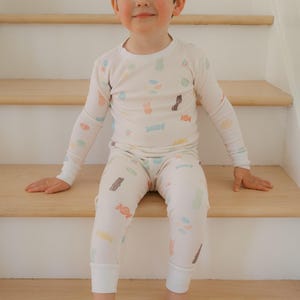Pode incluir: Uma crian&ccedil;a vestindo um conjunto de pijama creme com um padr&atilde;o de doces. A parte superior de manga comprida e as cal&ccedil;as apresentam ilustra&ccedil;&otilde;es coloridas de v&aacute;rios doces. A crian&ccedil;a est&aacute; sentada em escadas de madeira.