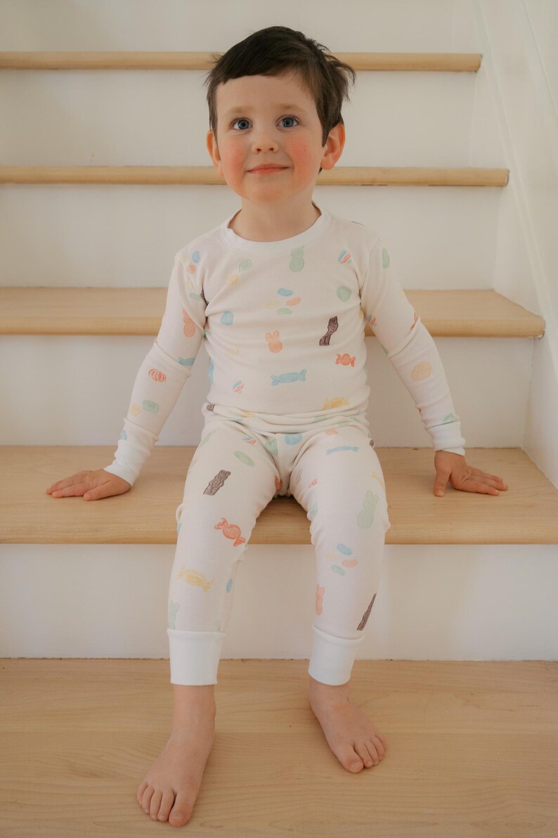 Pode incluir: Uma crian&ccedil;a vestindo um conjunto de pijama creme com um padr&atilde;o de doces. A parte superior de manga comprida e as cal&ccedil;as apresentam ilustra&ccedil;&otilde;es coloridas de v&aacute;rios doces. A crian&ccedil;a est&aacute; sentada em escadas de madeira.