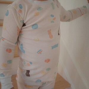 Pode incluir: Um conjunto de pijama de cor clara com um padr&atilde;o repetido de doces coloridos e ilustra&ccedil;&otilde;es de animais. A parte superior de manga comprida e as cal&ccedil;as t&ecirc;m punhos e acabamentos brancos. Os pijamas s&atilde;o projetados para crian&ccedil;as.