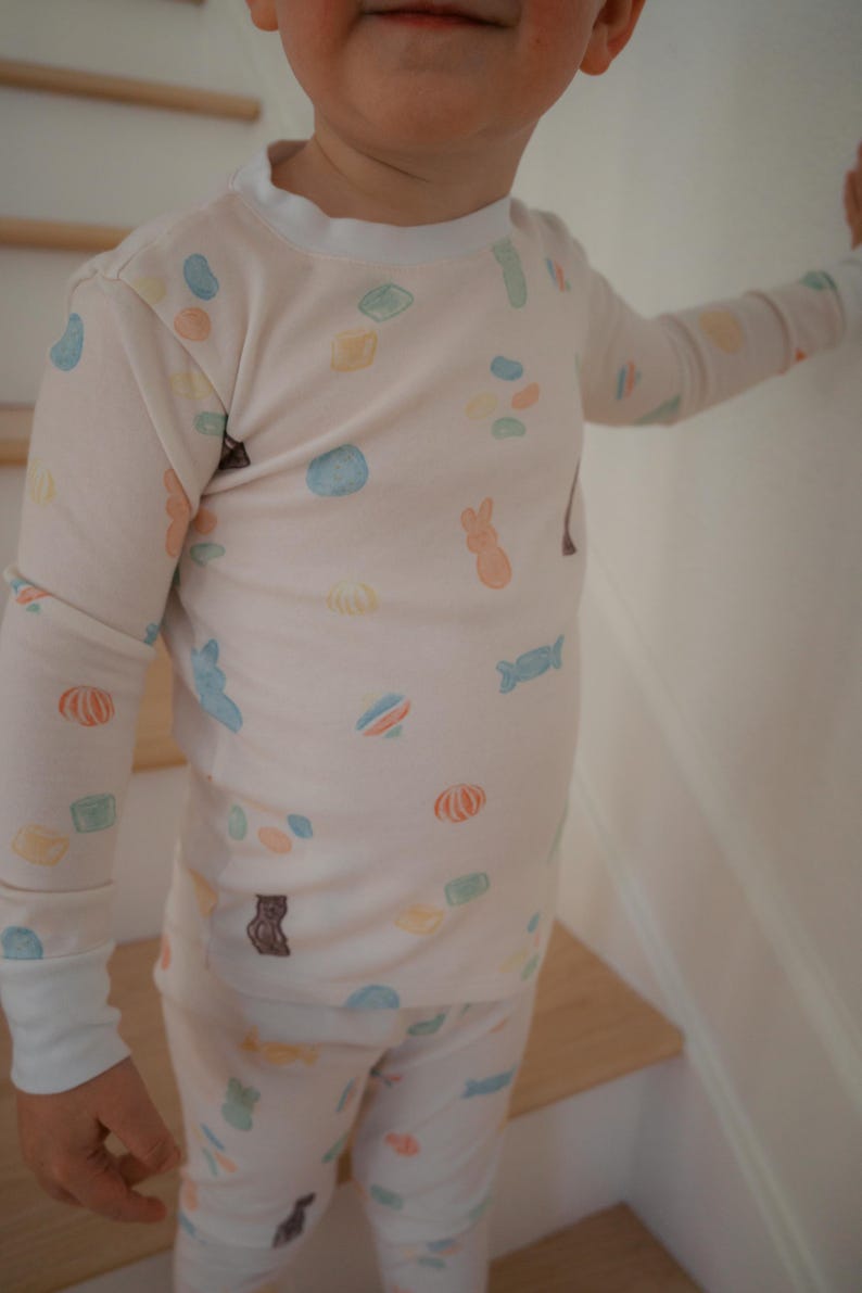 Pode incluir: Um conjunto de pijama de cor clara com um padr&atilde;o repetido de doces coloridos e ilustra&ccedil;&otilde;es de animais. A parte superior de manga comprida e as cal&ccedil;as t&ecirc;m punhos e acabamentos brancos. Os pijamas s&atilde;o projetados para crian&ccedil;as.