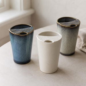 Könnte beinhalten: Drei Keramikbecher mit einem halbkreisförmigen oberen Rand, der ein fächerartiges Prägemuster aufweist. Ein Becher ist tiefblau mit dunklem Rand, einer ist mattweiß und der dritte ist graublau gesprenkelt mit einem dunklen, texturierten Rand.