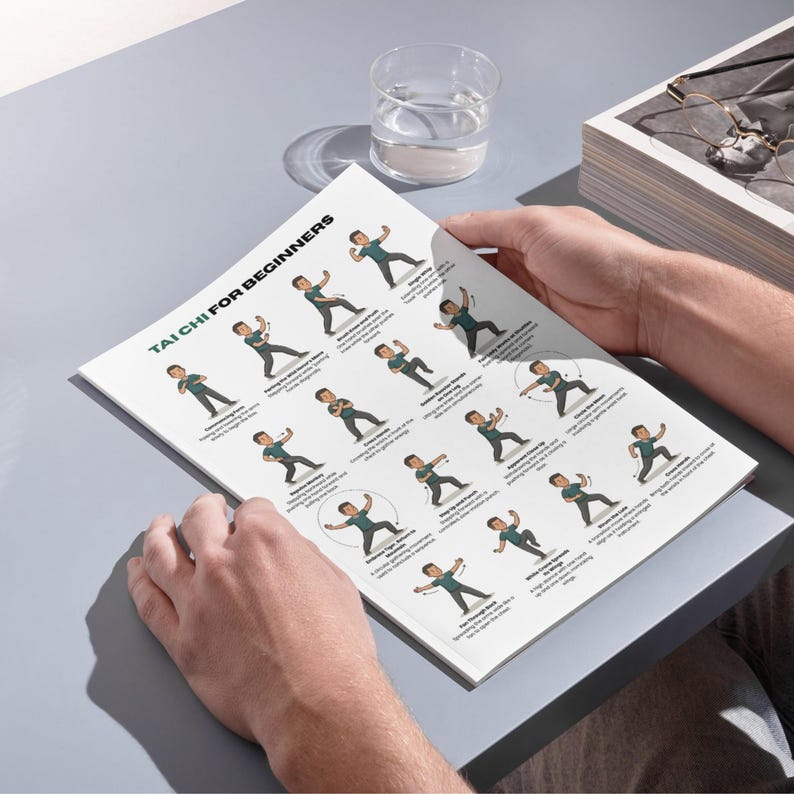 May include: A person holding a white booklet titled "TAI CHI FOR BEGINNERS." The booklet displays illustrated Tai Chi poses with accompanying text. A glass of water and a box with glasses are in the background. The booklet is on a gray surface.