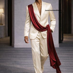 May include: A cream-colored satin suit with a draped burgundy sash. The suit features a two-button blazer and straight-leg trousers. The model is wearing cream-colored shoes. The overall look is elegant and formal.