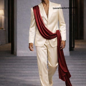 May include: A cream-colored suit with a draped burgundy sash. The suit features a two-button jacket and matching trousers. The model is wearing cream-colored shoes. The overall look is elegant and stylish.