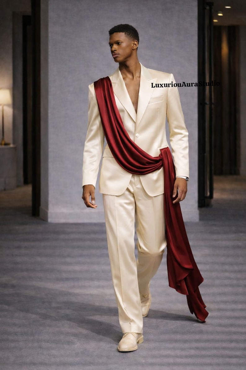 May include: A cream-colored suit with a draped burgundy sash. The suit features a two-button jacket and matching trousers. The model is wearing cream-colored shoes. The overall look is elegant and stylish.