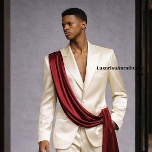 May include: A cream-colored suit with a maroon sash draped across the chest. The suit has a two-button closure and a notched lapel. The sash adds a pop of color and visual interest to the outfit. The text "LuxuriousAuraStudio" is visible.