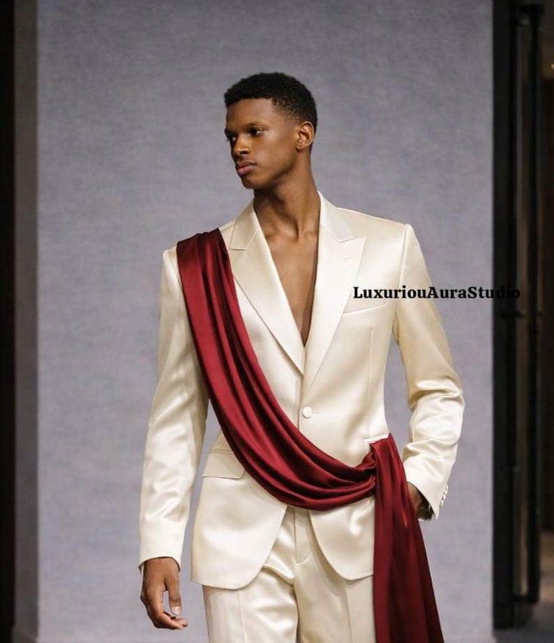 May include: A cream-colored suit with a maroon sash draped across the chest. The suit has a two-button closure and a notched lapel. The sash adds a pop of color and visual interest to the outfit. The text "LuxuriousAuraStudio" is visible.