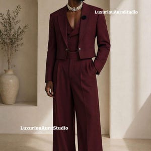 May include: A burgundy three-piece suit featuring a cropped jacket, vest, and wide-leg trousers. The suit has a tailored fit and is worn with a pearl necklace and a black flower pin. The outfit is a modern take on classic formal wear.