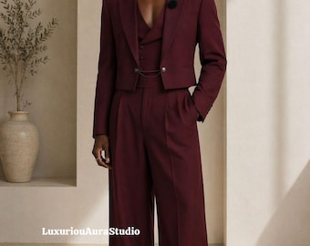 May include: A burgundy three-piece suit featuring a cropped jacket, vest, and wide-leg trousers. The suit has a tailored fit and is worn with a pearl necklace and a black flower pin. The outfit is a modern take on classic formal wear.