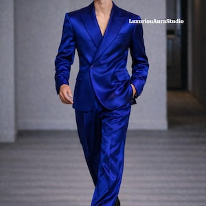 May include: A vibrant blue suit with a double-breasted jacket and matching trousers. The suit has a satin finish, reflecting light. The model is wearing black sunglasses and black dress shoes. The text "LuxuriousAuraStudio" is visible.