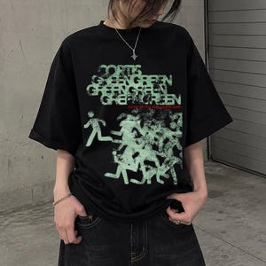 Könnte beinhalten: Ein schwarzes T-Shirt mit kurzen Ärmeln und einem hellgrünen Grafikdruck. Das Design zeigt das wiederholte Wort GREEN und eine Ansammlung stilisierter menschlicher Figuren in Bewegung. Der Text Cortis 26 Put your phone down ist rot gedruckt.