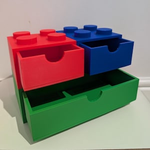 May include: A multi-coloured storage unit, designed to look like a building block toy. It has three pull-out drawers in red, blue, and green. The top of the unit has the characteristic raised studs of the toy.