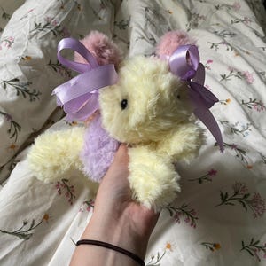 Könnte beinhalten: Ein Plüschtier in Form eines Hasen mit gelbem Fell und rosa und lila Akzenten. Der Hase hat rosa Bommelohren mit lavendelfarbenen Bändern. Das Spielzeug wird in einer Hand gehalten und liegt auf einem Bettlaken mit floralem Muster.