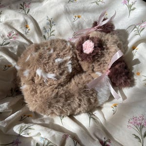 May include: A plush fawn toy with soft brown and white fur, featuring white spots on its back, a small pink crocheted flower on its head, and a light pink satin ribbon tied around its neck. The stuffed animal is curled in a resting position.