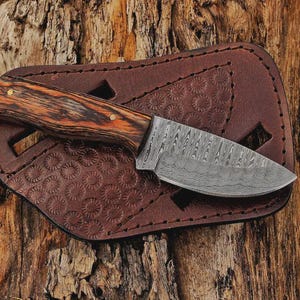 May include: A small, Damascus steel knife with a wooden handle and brass rivets rests on a brown leather sheath. The handle features a wood grain pattern in shades of brown and orange. The sheath has a decorative embossed pattern.