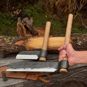 May include: Two hand-forged hatchets with wooden handles and dark metal blades. One hatchet is held by a person, while the other rests on a wooden surface. A leather sheath is also visible, suggesting outdoor or woodworking tools.