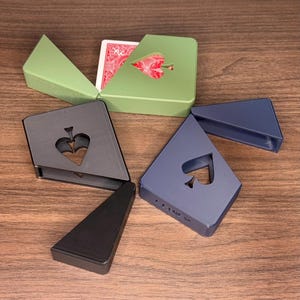 May include: A collection of playing card holders in various colors, including green, gray, blue, and black. Each holder features a spade-shaped cutout. One green holder contains a deck of cards with a red spade design.