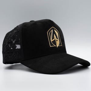 Puede incluir: Gorra de béisbol negra con un emblema dorado en la parte delantera y una parte trasera de malla de encaje negro. La gorra tiene una visera curva y una corona estructurada. El emblema dorado es un diseño estilizado.