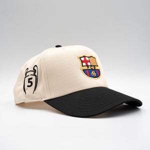 May include: A beige baseball cap with a black brim. The cap features an embroidered FC Barcelona logo in red, blue, and yellow. On the side is a black outline of a trophy with the number 5.