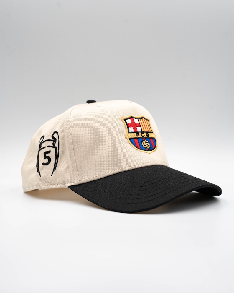 May include: A beige baseball cap with a black brim. The cap features an embroidered FC Barcelona logo in red, blue, and yellow. On the side is a black outline of a trophy with the number 5.