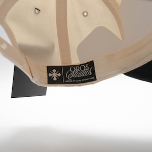 May include: Cream-colored baseball cap with a black brim. The underside of the cap features a cream-colored fabric and a black tag with the text "OROS Studios" and a graphic. The cap has a classic six-panel design.