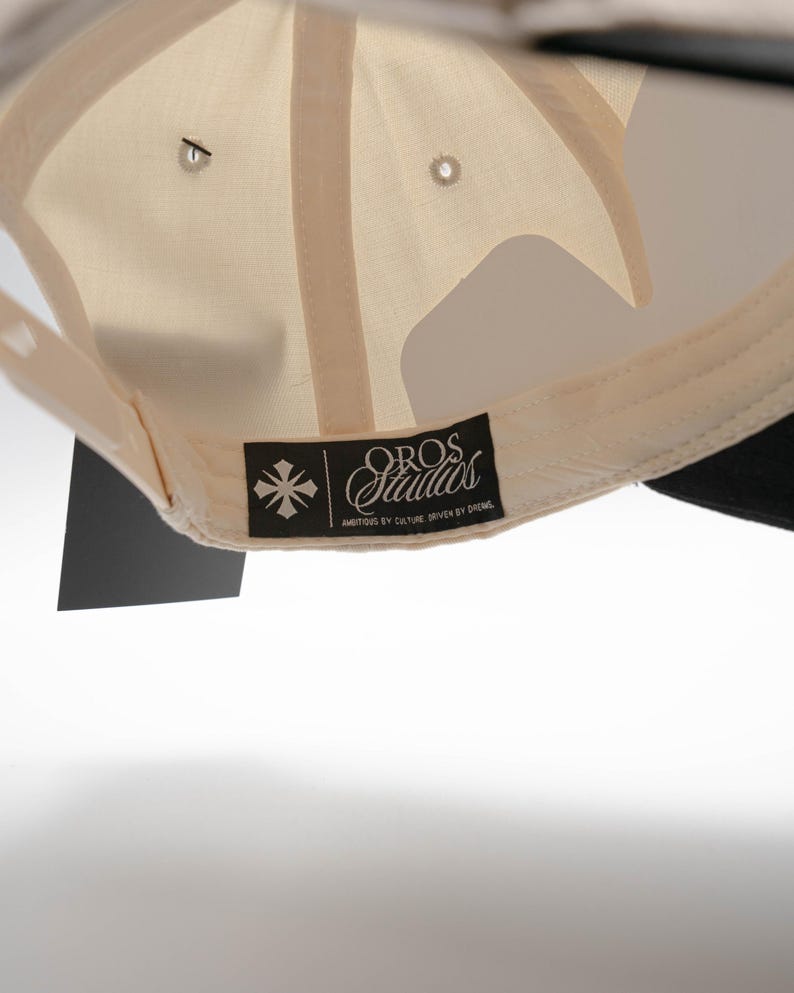 May include: Cream-colored baseball cap with a black brim. The underside of the cap features a cream-colored fabric and a black tag with the text "OROS Studios" and a graphic. The cap has a classic six-panel design.