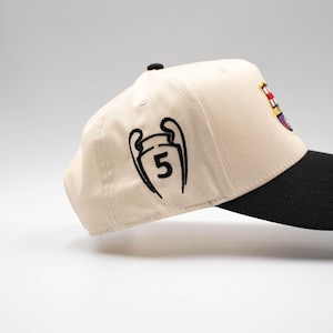 May include: A beige baseball cap with a black brim. The cap features a black embroidered design of a trophy with the number 5, and a team logo on the side. The cap is a sports fan accessory.