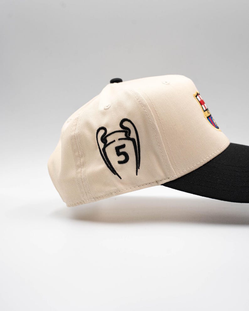 May include: A beige baseball cap with a black brim. The cap features a black embroidered design of a trophy with the number 5, and a team logo on the side. The cap is a sports fan accessory.