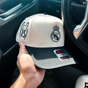 Może przedstawiać: Beżowo-czapka z daszkiem z herbem Realu Madryt i numerem 15 wyhaftowanym z boku. Czapka ma płaski daszek i klasyczny krój.