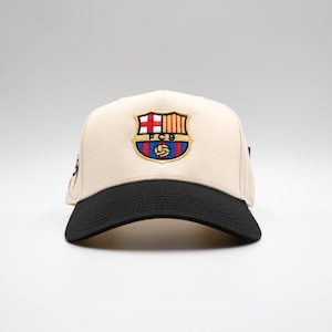 May include: A beige baseball cap with a black brim. The cap features an embroidered emblem with the letters "FCB" and a soccer ball. The emblem is in red, blue, yellow, and white.