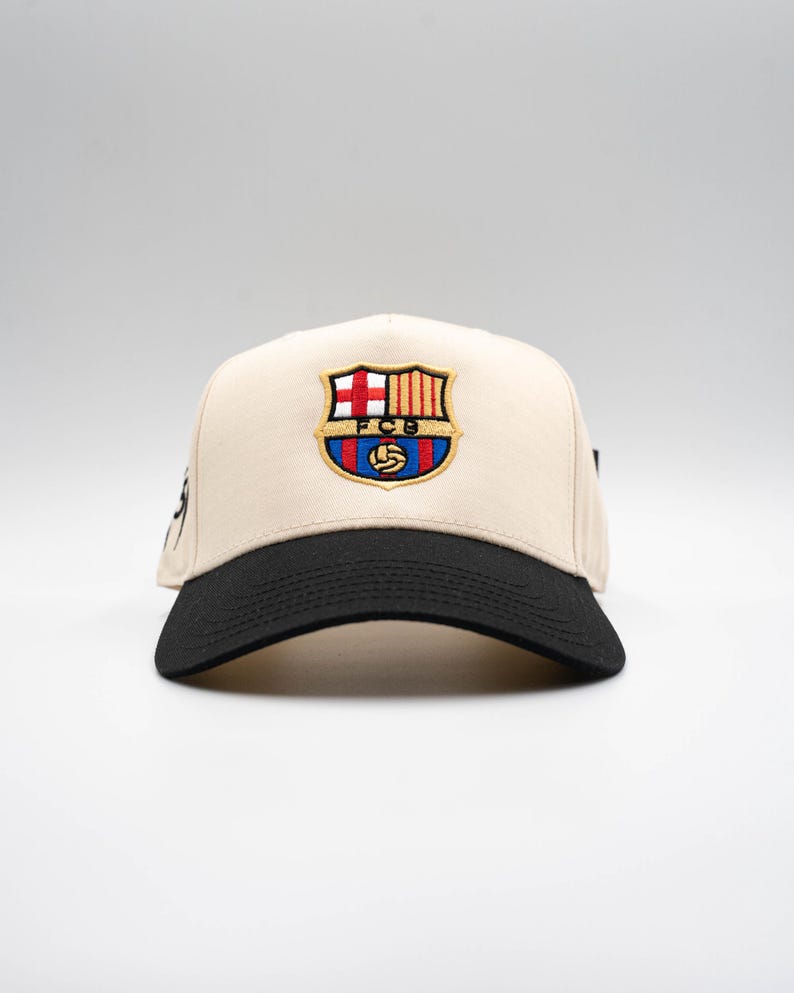 May include: A beige baseball cap with a black brim. The cap features an embroidered emblem with the letters "FCB" and a soccer ball. The emblem is in red, blue, yellow, and white.