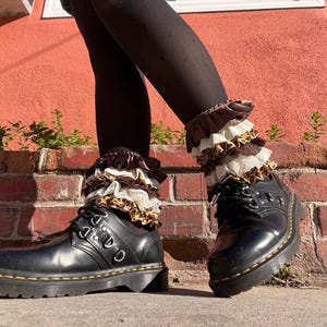 May include: Black leather boots with a lace-up design and a thick sole. The boots are paired with ruffled ankle cuffs in brown, white, and leopard print patterns. The background features a red brick wall.