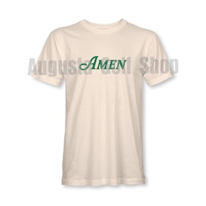 May include: A cream-coloured t-shirt featuring the word "AMEN" in green script across the chest. The text "Augusta Golf Shop" is faintly visible above the shirt. The shirt has short sleeves and a crew neck.
