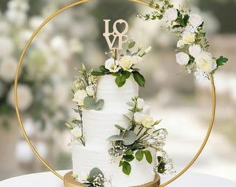 May include: A three-tiered white wedding cake decorated with white flowers and greenery. The cake is on a wooden base and adorned with a gold hoop and a wooden "LOVE" cake topper. The background is blurred, suggesting an outdoor setting.