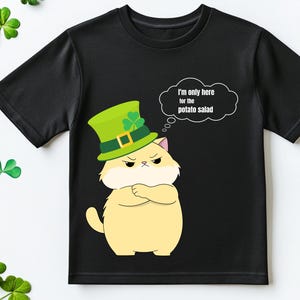 Op de afbeelding: Zwart T-shirt met een cartoonkat met een groene St. Patrick's Day hoed. De kat heeft een humeurige uitdrukking en gekruiste armen. Een tekstballon zegt: "Ik ben hier alleen voor de aardappelsalade."