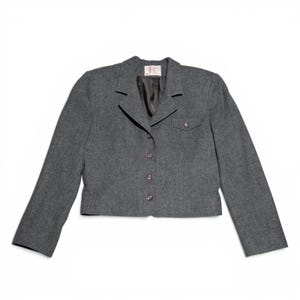 May include: A gray blazer with a notched lapel, three buttons, and a small pocket. The cropped jacket has long sleeves and a dark lining. The blazer is made of a textured fabric and is displayed on a white background.