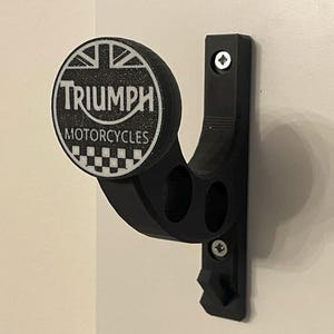 Peut inclure: Un crochet mural noir pour casque doté d'un emblème circulaire avec le texte blanc Triumph Motorcycles, un motif Union Jack et un damier. Le crochet possède un bras de support incurvé et est fixé au mur avec deux vis visibles.