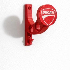 Può includere: Un portachiavi rosso da parete con il logo Ducati. Il supporto ha un gancio e un design circolare con l'emblema Ducati in bianco su sfondo rosso. L'articolo è montato su una parete bianca.
