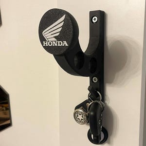 Peut inclure: Un crochet mural noir pour casque avec un embout circulaire arborant le logo ailé Honda et le texte en blanc. Le support présente un bras incurvé avec deux découpes circulaires et une patère inférieure pour les clés, fixé par deux vis.