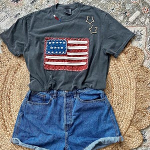 May include: A dark gray t-shirt with a sewn-on American flag design, paired with blue denim shorts. The flag features red and white stripes and a blue square with white stars. Two wooden star-shaped embellishments are on the shirt.