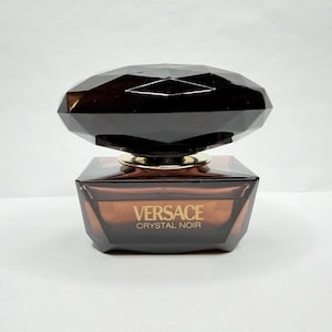 May include: A Versace Crystal Noir perfume bottle. The bottle is a dark brown color with a faceted, gem-like cap. The perfume bottle has a rectangular shape with the words "Versace Crystal Noir" printed on the front.
