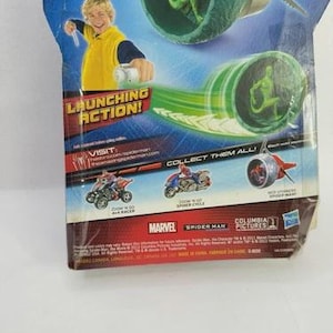May include: A Spider-Man action figure toy package. The package features a green and silver projectile launcher with a green figure inside. The text "LAUNCHING ACTION!" is visible, along with the Marvel and Spider-Man logos.