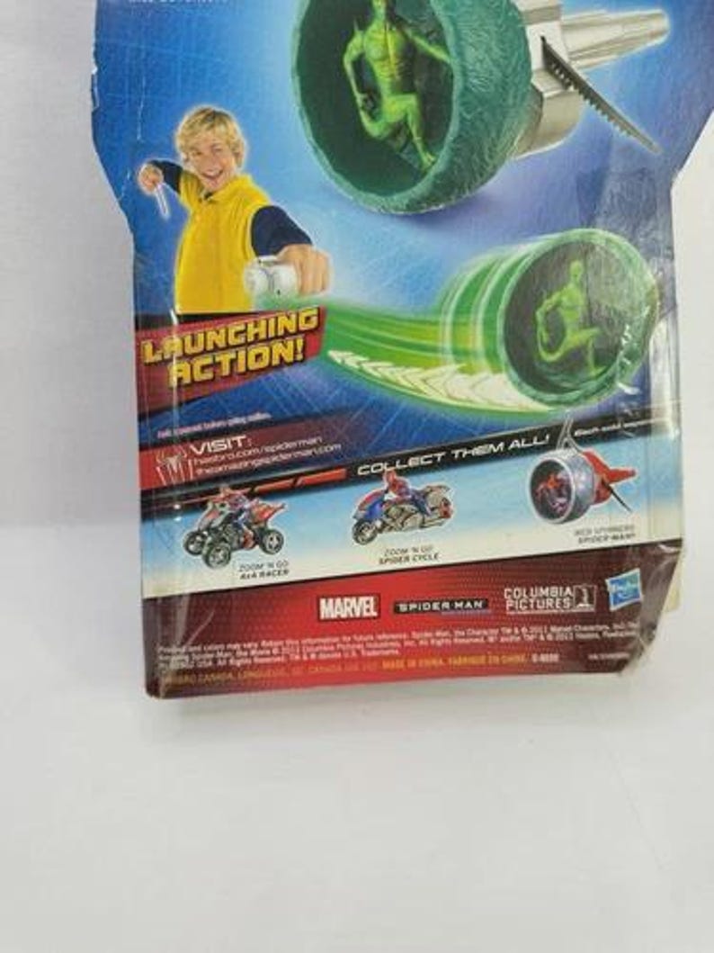 May include: A Spider-Man action figure toy package. The package features a green and silver projectile launcher with a green figure inside. The text "LAUNCHING ACTION!" is visible, along with the Marvel and Spider-Man logos.