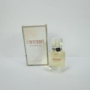 Puede incluir: Un frasco de perfume Givenchy L'Interdit Eau de Toilette con su caja. El frasco transparente tiene un tapón plateado y una etiqueta roja. La caja blanca muestra el nombre y el logotipo del perfume.