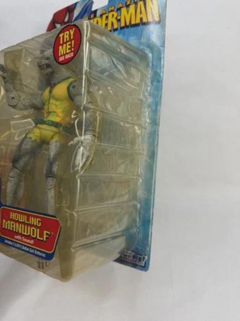 May include: A Howling Manwolf action figure from The Amazing Spider-Man series, packaged in a clear plastic case. The figure is gray and yellow, with the text "TRY ME!" and "HOWLING MANWOLF" on the packaging.