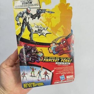 May include: A packaged Venom action figure from the "Toxic Blast" series. The figure is in a black and white suit, holding a weapon. The packaging is yellow and red, with the text "Web Missile!" and "Help Spider-Man Battle His Fiercest Foes!"