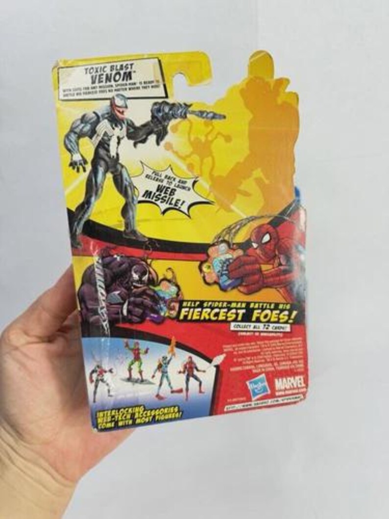 May include: A packaged Venom action figure from the "Toxic Blast" series. The figure is in a black and white suit, holding a weapon. The packaging is yellow and red, with the text "Web Missile!" and "Help Spider-Man Battle His Fiercest Foes!"
