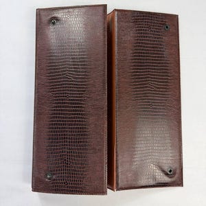 May include: Two brown, rectangular leather-like cases with a reptile skin texture. Each case has a dark brown exterior and a lighter brown interior. The cases are secured with a single, dark-colored button on each. The cases are open, revealing the interior.